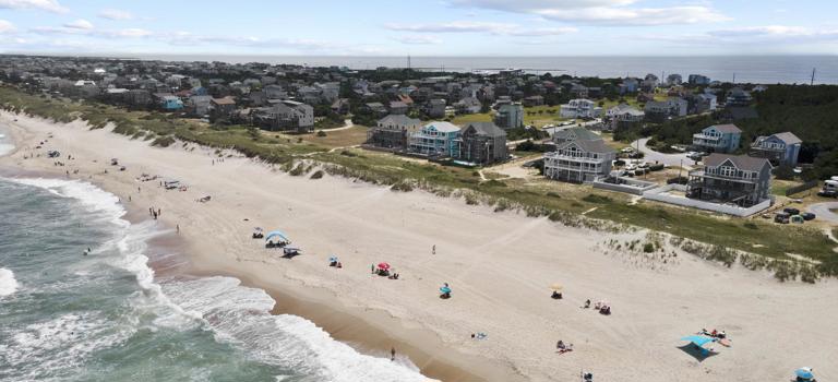 A wide sandy beach in Waves, NC as seen from a drone over the ocean looking SW to vacation homes and the horizon